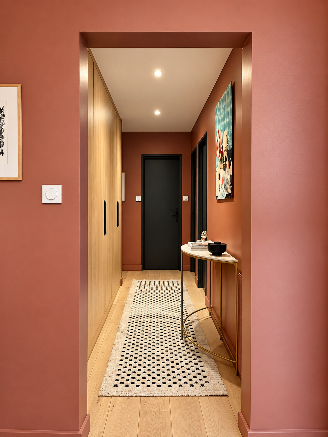 Couloir contemporain aux murs terracotta, avec placards sur mesure en bois clair, portes noires, console élégante en laiton et tapis graphique noir et blanc.