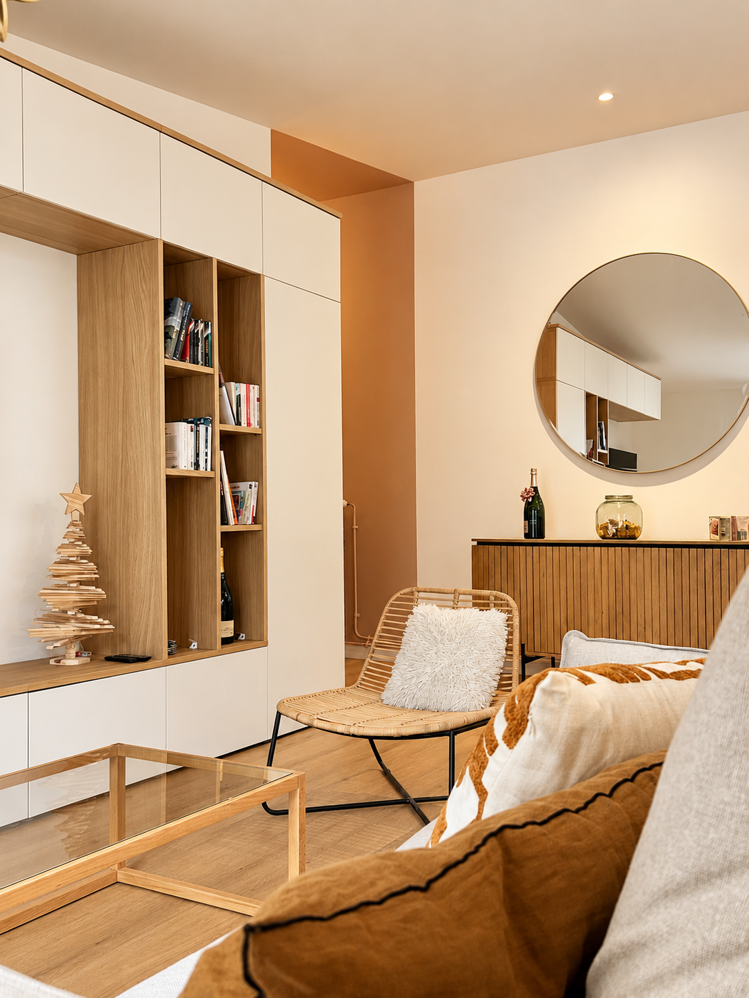 Salon contemporain et chaleureux avec un grand meuble TV sur mesure associant façades blanches et bois naturel. Des niches ouvertes accueillent des livres et des objets décoratifs, tandis qu’un fauteuil en rotin avec coussin blanc apporte une touche bohème. Le parquet en bois clair et l’éclairage chaleureux complètent cet aménagement élégant et fonctionnel.