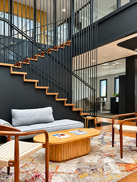 Espace détente au pied d’un escalier en bois et métal noir, avec mobilier chaleureux, tapis coloré et cloisons vitrées