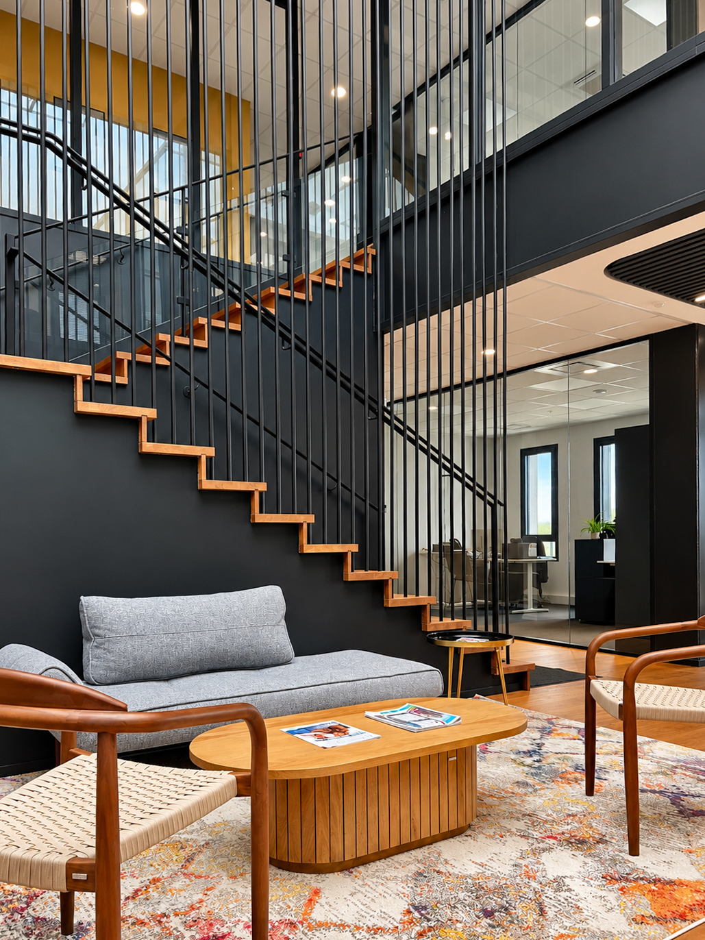 Espace détente au pied d’un escalier en bois et métal noir, avec mobilier chaleureux, tapis coloré et cloisons vitrées