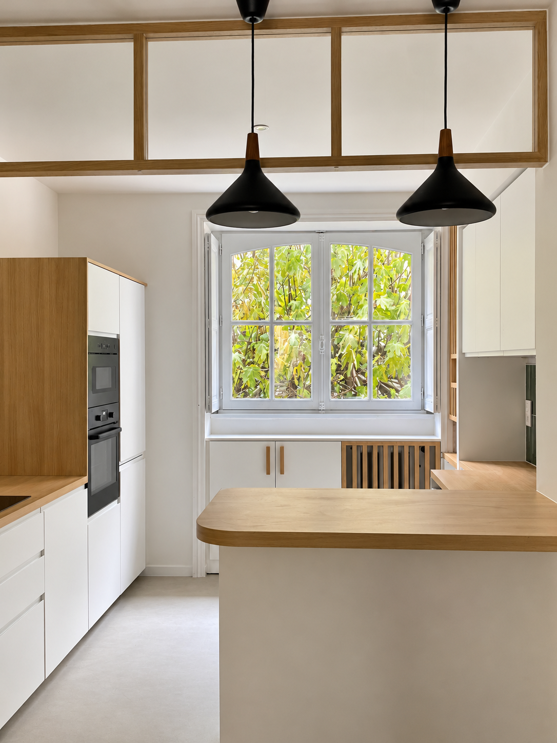 Cuisine contemporaine et lumineuse aux lignes épurées, associant des meubles blancs à des éléments en bois naturel clair. Une élégante verrière intérieure en bois structure l’espace tout en laissant circuler la lumière. Les plans de travail en bois, la crédence en carrelage blanc brillant et les équipements noirs, notamment la hotte inclinée et la plaque de cuisson, créent un contraste harmonieux. Une suspension noire et un sol beige clair complètent cet aménagement moderne, chaleureux et minimaliste.