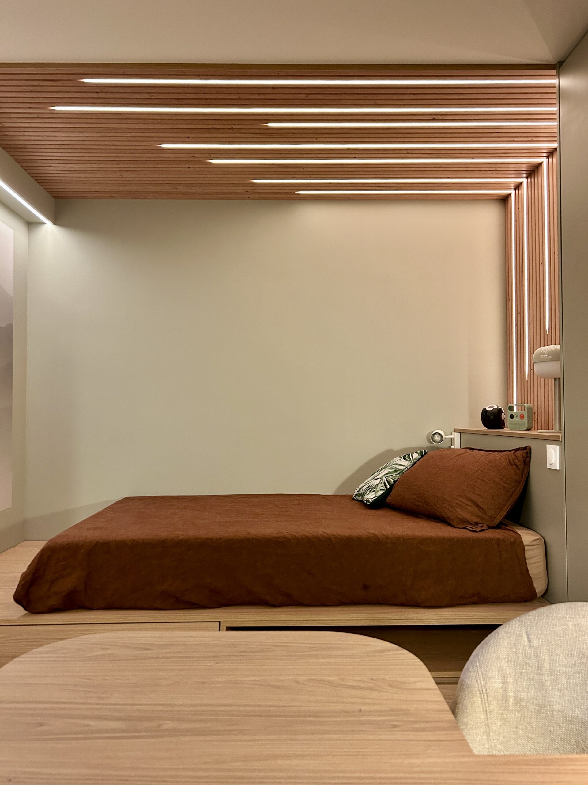 Aménagement sur mesure d’une chambre avec lit sur estrade en bois clair, alcôve habillée de tasseaux de bois et éclairage LED intégré au plafond et au mur, dans un style contemporain et chaleureux.